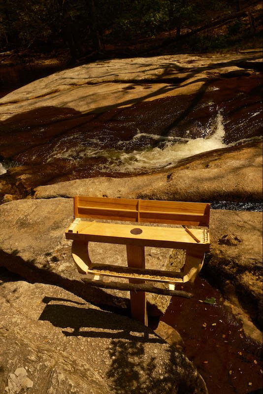 The sculpture seen from above on granite beside a stream, lid open, seed in the tray.