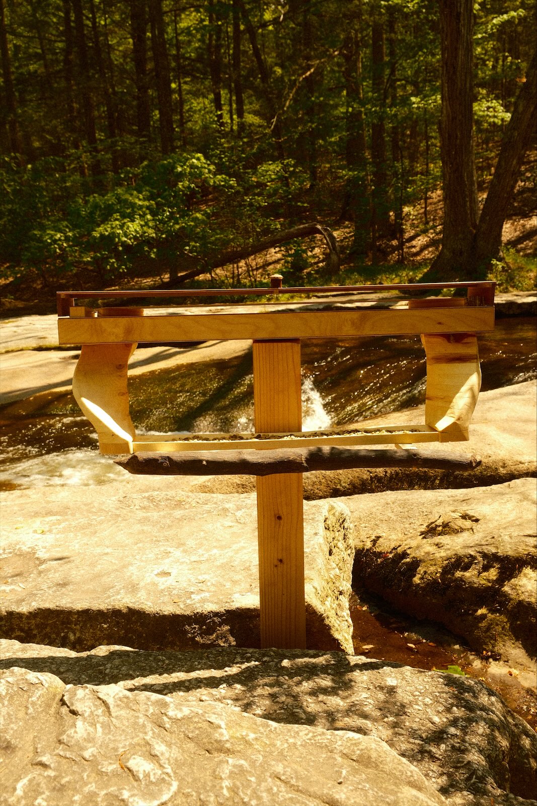 The sculpture standing in a cleft of granite beside a stream, forest behind, seen straight on.