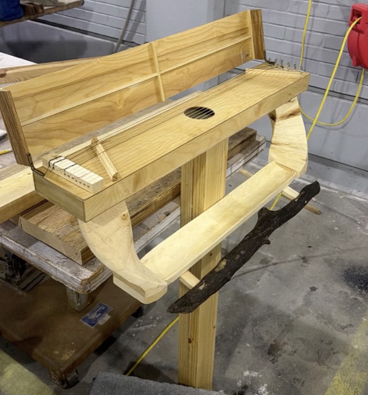 In the workshop: the assembled sculpture on its post, lid open to show the strings and sound hole, carved supports holding the tray, and the branch perch fixed in front.