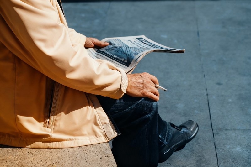 A seated reader in a pale jacket holds a newspaper and a cigarette above blue-gray pavement.