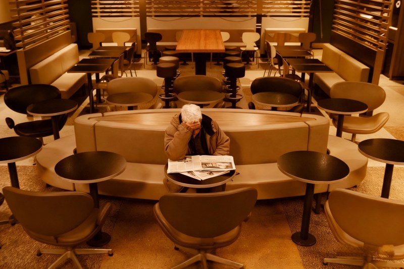 A lone newspaper reader sits with a hand over their face among rows of empty café tables.