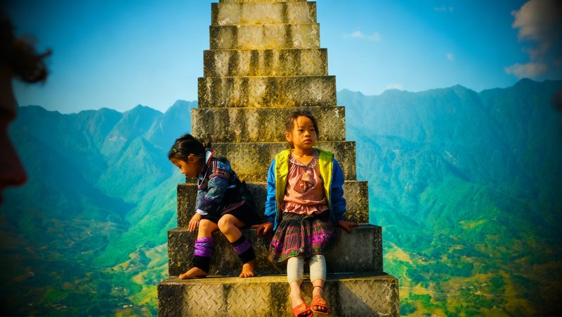 Two young girls in brightly patterned clothing sit on the steps of a tall concrete stairway, one looking down to the side and one toward the camera, with green mountain slopes and blue sky behind and the blurred edge of a face at the left of the frame.